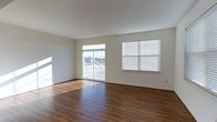 This three-floor space offers a clean, modern aesthetic defined by crisp white walls and rich, dark hardwood-style flooring that runs throughout. The main living area is generously proportioned, featuring an open layout that allows natural light to pour in from multiple sources. A sliding glass door leads directly to a private balcony, offering outdoor views and additional brightness that enhances the airy feel of the room. A adjacent window fitted with horizontal blinds provides further light control and ventilation options.

The flooring makes a strong visual statement, with warm, dark tones that contrast effectively against the neutral white walls and ceiling, creating a grounded yet bright atmosphere. The ceiling height adds to the sense of openness, contributing to the overall spacious feel of the living area.

The space maintains a neutral, versatile palette throughout, making it well-suited for a variety of interior styles. Spread across three floors, the layout provides ample opportunity for dedicated living, sleeping, and functional zones. The combination of natural light, outdoor access, and clean architectural lines gives this space a welcoming and practical character.