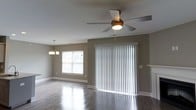 This single-floor residence offers a thoughtfully designed layout centered around a spacious, open living area. The room features rich, dark hardwood-style flooring that runs throughout, providing a sleek and polished aesthetic that complements the neutral gray wall tones. A five-blade ceiling fan with an integrated light fixture is centrally mounted, ensuring comfortable airflow and supplemental lighting across the room. A large sliding glass door, dressed with white vertical blinds, serves as a prominent architectural feature, allowing generous natural light to flood the interior while offering potential access to an outdoor area. A secondary window along the adjacent wall further enhances the bright, airy atmosphere of the room. An elegant chandelier-style pendant light is positioned toward the dining area, adding a refined touch to the open-plan space. Clean white baseboards frame the room, and a partial view of a fireplace mantle is visible toward one corner, hinting at additional character within the home. The overall ambiance is clean, modern, and move-in ready, making it a welcoming and functional living environment suited for a variety of lifestyles.