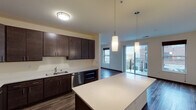 This single-floor unit showcases a thoughtfully designed open-concept layout, with the kitchen and living area flowing seamlessly together. The kitchen is a standout feature, outfitted with rich dark espresso cabinetry extending to the ceiling for ample storage, light-toned quartz countertops, and a full suite of stainless steel appliances including a refrigerator, range, dishwasher, and built-in microwave. A spacious island with pendant lighting provides additional prep space and visual separation between the kitchen and living area. The living room is bathed in natural light from multiple windows and a sliding glass door that opens to a private balcony with outdoor views. Dark hardwood-style flooring runs throughout the main living spaces, adding warmth and continuity. The bedrooms feature plush carpeting and large windows that bring in natural light. Both bathrooms are finished with dark wood vanities, light stone-toned countertops, large mirrors, and hardwood-style flooring. One bathroom includes accessible grab bars and a walk-in shower, while the second features a full tub-shower combination. The unit's neutral color palette and consistent cabinetry finishes create a cohesive, modern aesthetic throughout.
