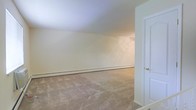This single-floor space presents a straightforward and functional layout with a clean, move-in-ready aesthetic. The main room is finished with wall-to-wall carpeting in a neutral beige tone, providing a comfortable underfoot feel while complementing the cream-colored walls throughout. The room offers a generous open floor plan, providing flexibility for a variety of furniture arrangements and uses.

A baseboard heating system runs along the base of the main wall, ensuring efficient and even heat distribution throughout the space. The smooth, light-colored ceiling reflects ambient light well, contributing to a bright and airy atmosphere despite the absence of large windows visible in this view.

A white panel door with a brushed nickel hardware finish is positioned to one side of the room, providing access to an adjacent area. The overall color palette — consisting of soft whites, creams, and warm beiges — creates a cohesive and calm environment. The space is currently unoccupied, allowing prospective occupants to clearly envision their own design and layout preferences. Its understated finishes and open arrangement make it a versatile and practical living space.