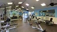 This single-floor fitness center is thoughtfully organized into distinct workout zones, offering a comprehensive exercise environment. The strength training area features rubberized flooring and is outfitted with a full rack of dumbbells, adjustable benches, and a variety of weight machines, all positioned alongside floor-to-ceiling mirrors that enhance the sense of space and allow users to monitor their form. Ceiling fans mounted throughout provide consistent airflow, complementing the bright, recessed panel lighting overhead.

Adjacent to the strength area, a dedicated cardio zone houses a row of treadmills and additional cardio machines, positioned near large windows that invite natural light into the space. A separate room with hardwood flooring serves as a group exercise or stretching studio, offering a clean, open area suitable for yoga, aerobics, or other fitness classes.

The walls are painted in a soft blue tone with white wainscoting accents, creating a clean and motivating atmosphere. Instructional fitness posters are displayed throughout, adding an informative and professional touch to the overall environment.