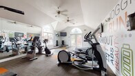 This single-level fitness center is designed to inspire and support a comprehensive workout experience. The space is defined by its striking vaulted ceilings, which lend an open, airy feel, while dual ceiling fans ensure comfortable airflow throughout. Large arched windows and glass double doors flood the interior with abundant natural light and offer pleasant views of lush, tree-lined surroundings, creating an inviting atmosphere for exercise.

The cardio section is well-appointed with multiple treadmills, elliptical trainers, and stationary bikes — including both upright and recumbent models — catering to a variety of fitness preferences. A Life Fitness cable machine anchors the strength training area, complemented by a full dumbbell rack and an adjustable weight bench. Wall-mounted flat-screen televisions are positioned throughout for entertainment during workouts.

The interior is further elevated by vibrant, fitness-themed wall murals featuring motivational typography and bold graphic illustrations of athletes in motion, adding an energetic and dynamic character to the space. Full-length mirrors expand the sense of space and allow for proper form checks during exercise. The flooring combines durable carpet tile with accent-colored gym mats in orange and yellow, adding both function and visual interest. This fitness center delivers a well-rounded, thoughtfully designed environment for all levels of exercise.