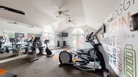 This single-level fitness center is designed to inspire and support a comprehensive workout experience. The space is defined by its striking vaulted ceilings, which lend an open, airy feel, while dual ceiling fans ensure comfortable airflow throughout. Large arched windows and glass double doors flood the interior with abundant natural light and offer pleasant views of lush, tree-lined surroundings, creating an inviting atmosphere for exercise.

The cardio section is well-appointed with multiple treadmills, elliptical trainers, and stationary bikes — including both upright and recumbent models — catering to a variety of fitness preferences. A Life Fitness cable machine anchors the strength training area, complemented by a full dumbbell rack and an adjustable weight bench. Wall-mounted flat-screen televisions are positioned throughout for entertainment during workouts.

The interior is further elevated by vibrant, fitness-themed wall murals featuring motivational typography and bold graphic illustrations of athletes in motion, adding an energetic and dynamic character to the space. Full-length mirrors expand the sense of space and allow for proper form checks during exercise. The flooring combines durable carpet tile with accent-colored gym mats in orange and yellow, adding both function and visual interest. This fitness center delivers a well-rounded, thoughtfully designed environment for all levels of exercise.