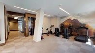 Located at 4901 Montgomery Lane in Bethesda, Maryland, this well-appointed fitness center offers a comprehensive workout environment designed for convenience and comfort. The space is thoughtfully laid out on a single level, with neutral-toned checkerboard carpet flooring and sleek recessed linear lighting that runs along the ceiling, creating a clean, modern ambiance throughout.

The cardio zone is well-equipped with multiple treadmills, elliptical cross-trainers, and stationary cycling bikes, all featuring integrated digital displays. A dramatic floor-to-ceiling mountain landscape mural serves as a bold focal point, energizing the workout environment. Large windows along one wall invite abundant natural light and provide views of the surrounding Bethesda streetscape.

The functional training area includes a rack of medicine balls in varying weights, stability/exercise balls on a dedicated stand, a set of dumbbells, and kettle bells — catering to a wide range of strength and conditioning exercises. Full-length mirrors on one wall enhance the sense of space and allow for proper form monitoring.

A dedicated amenity nook features built-in wood-accented shelving stocked with fresh rolled towels, a water dispenser, and a waste station, ensuring members have everything needed for a comfortable session. The overall design balances practicality with an upscale aesthetic befitting its Bethesda location.
