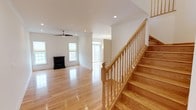 This thoughtfully designed 3-floor home offers a seamless blend of classic charm and functional living. The main level immediately impresses with gleaming hardwood floors that extend throughout the open living area, reflecting abundant natural light that streams in through multiple windows fitted with cellular shades. A ceiling fan with a sleek profile anchors the spacious living room, which also features a freestanding fireplace with a black finish — a striking focal point ideal for cooler months. Recessed lighting adds a polished, modern touch to the otherwise clean white walls and ceiling. A beautifully crafted wood staircase with turned balusters and a smooth handrail serves as both a functional and architectural centerpiece, connecting all three floors in style. The flow of the main level extends toward what appears to be additional rooms or transitional spaces, hinting at a generous and versatile floor plan. With its warm wood tones, neutral palette, and well-proportioned rooms, this home exudes a bright and welcoming ambiance suited for comfortable everyday living across all three levels.