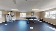 This single-floor fitness facility is thoughtfully designed to accommodate a variety of workout styles and user needs. The main exercise area is spacious and open, finished with durable rubber flooring that covers a generous workout zone ideal for strength training and functional movement. A multi-tiered dumbbell rack lines the wall and is accompanied by adjustable workout benches, a multi-function cable and pull-up station, and a stability ball, providing ample options for resistance training. Ceiling fans keep the space comfortable, while fluorescent lighting and multiple windows along the walls allow natural light to brighten the interior and offer outdoor views of the surrounding area. The cardio section features Nautilus treadmills positioned near a wall-mounted flat-screen television, making workouts more engaging. A convenient beverage vending machine is situated near the entrance, and glass-paned double doors provide easy access to the exterior. A separate, carpeted children's play room is also part of the facility, decorated with colorful wall art and furnished with soft toys and a bean bag chair, offering a welcoming space for younger visitors. Textured ceilings and crown molding throughout add a finished, polished look to the overall environment.