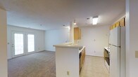 This single-floor residence is designed with an open, functional layout that promotes ease of movement and a cohesive flow between living spaces. The living area is finished with wall-to-wall carpet in a warm neutral tone, providing a comfortable underfoot feel, while white walls and a textured ceiling contribute to the bright, clean aesthetic throughout. A full-lite exterior door with blinds allows natural light to filter into the main living space, enhancing its open character.

The kitchen is positioned at the heart of the home and features a peninsula-style breakfast bar that visually and physically connects the kitchen to the living area. Warm wood-toned cabinetry provides ample storage, while tile flooring in the kitchen zone offers easy maintenance. A range is included among the appliances. Recessed and overhead lighting illuminate the kitchen workspace effectively.

A hallway visible from the main living area leads to the additional rooms of the residence, suggesting a well-organized floor plan with clearly defined private and shared spaces. Throughout the home, neutral tones, consistent finishes, and practical design choices create a versatile environment well-suited to a variety of living arrangements. The overall ambiance is light, clean, and move-in ready.