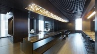 This elegantly designed single-floor space exudes a refined, contemporary aesthetic that balances dramatic visual impact with functional sophistication. At the heart of the space stands a commanding reception or communal counter finished in richly veined marble, anchored by sleek dark wood paneling that extends throughout the interior, lending a sense of warmth and gravitas. Overhead, a large rectangular ceiling installation illuminated with sculptural branch-and-leaf motifs serves as a stunning focal point, casting a warm, organic glow against the dark grid ceiling above. The flooring transitions from rich dark wood to lighter wood-toned planks, subtly defining zones within the open layout. Toward the rear, a built-in shelving unit lined with books and objects creates an inviting library-style backdrop, flanked by modern white swivel lounge chairs that introduce a clean contrast to the deeper tones of the room. Natural light filters in through floor-to-ceiling windows, brightening the lounge area and balancing the moody, atmospheric interior lighting. Minimalist benches offer additional seating and reinforce the space's clean-lined design language. Overall, the space conveys a polished, upscale ambiance well-suited for a corporate lobby, boutique hotel, or premium commercial venue.