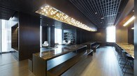 This elegantly designed single-floor space exudes a refined, contemporary aesthetic that balances dramatic visual impact with functional sophistication. At the heart of the space stands a commanding reception or communal counter finished in richly veined marble, anchored by sleek dark wood paneling that extends throughout the interior, lending a sense of warmth and gravitas. Overhead, a large rectangular ceiling installation illuminated with sculptural branch-and-leaf motifs serves as a stunning focal point, casting a warm, organic glow against the dark grid ceiling above. The flooring transitions from rich dark wood to lighter wood-toned planks, subtly defining zones within the open layout. Toward the rear, a built-in shelving unit lined with books and objects creates an inviting library-style backdrop, flanked by modern white swivel lounge chairs that introduce a clean contrast to the deeper tones of the room. Natural light filters in through floor-to-ceiling windows, brightening the lounge area and balancing the moody, atmospheric interior lighting. Minimalist benches offer additional seating and reinforce the space's clean-lined design language. Overall, the space conveys a polished, upscale ambiance well-suited for a corporate lobby, boutique hotel, or premium commercial venue.