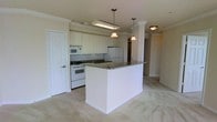 This single-floor residence offers a thoughtfully designed open-concept layout that connects the kitchen and living areas in a seamless, functional arrangement. The kitchen is outfitted with crisp white cabinetry, dark granite countertops, and a tile backsplash that adds visual texture to the space. A central peninsula with matching granite provides additional prep space and serves as a natural divider between the kitchen and the adjoining living area. White appliances, including a refrigerator, range, and built-in microwave, are neatly integrated into the cabinetry. Pendant lighting hangs above the peninsula, casting warm, focused light over the workspace. Decorative columns frame the transition between the kitchen and the open living space, lending an architectural elegance to the layout. The living area features soft, neutral carpeting that contrasts with the tile flooring in the kitchen, creating a clear and comfortable zone distinction. Crown molding runs along the ceiling throughout, adding a refined, polished character to the interior. The overall ambiance is clean, bright, and inviting, with a neutral palette that provides a versatile backdrop suited to a variety of interior styles.