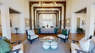 This refined single-floor community or amenity space showcases an impressive architectural design defined by dramatic cathedral-height ceilings, bold exposed timber beam frameworks, and warm hardwood flooring throughout. The grand central lobby greets visitors with a striking arrangement of elegant armchairs, a sculptural glass-top coffee table, and a pair of statement chandeliers suspended beneath the soaring ceiling. Large clerestory windows and full-height glass doors flood the space with natural light while offering views of the outdoor surroundings.

A welcoming reception counter finished in soft sage green with a marble top anchors the main hall, which flows seamlessly into multiple distinct gathering areas. One lounge features richly upholstered leather seating centered around a stone-clad fireplace with a herringbone tile surround and a dark wood ceiling, creating an intimate and cozy ambiance. Another casual lounge area opens directly to the outdoors through French doors, bathed in sunlight and furnished with comfortable armchairs and coordinating area rugs.

A dedicated conference room offers a polished oval wood table surrounded by tufted upholstered chairs, providing a functional and professional setting. Throughout the space, warm wall sconces, textured wainscoting, curated greenery, and thoughtful décor details contribute to an atmosphere that is both welcoming and sophisticated.