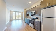 This well-appointed single-floor residence showcases a thoughtfully designed open-concept layout that seamlessly connects the kitchen and living areas. The contemporary kitchen is a standout feature, outfitted with rich dark wood cabinetry extending floor-to-ceiling, granite countertops, a subway tile backsplash, and a full suite of stainless steel appliances including a refrigerator, electric range, built-in microwave, and dishwasher. Light wood-style flooring runs throughout the unit, lending warmth and continuity to every room. The living area benefits from an abundance of natural light courtesy of a striking bank of tall windows, creating an airy, open atmosphere with outdoor views. The bedrooms are bright and well-proportioned, with one featuring a ceiling fan and a large window overlooking the exterior. A walk-in closet with wire shelving provides generous storage. The bathroom is finished with clean, modern materials including large-format tile flooring, full subway tile surround at the tub/shower, a granite-topped vanity with dark wood cabinetry, and brushed nickel fixtures. Neutral wall tones and recessed lighting throughout complete the polished, contemporary feel of this well-designed space.