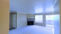 This single-floor space offers a welcoming and functional layout designed for comfortable living. The main living area is a standout feature, showcasing a tile-surround fireplace with a classic mantle that serves as a natural focal point of the room. Plush wall-to-wall carpeting adds warmth underfoot, while crisp white crown molding along the ceiling provides an elegant finishing touch against the neutral, warm-toned walls. Natural light flows generously into the space through a large picture window and an adjacent sliding glass door, which also provides access to an outdoor area, creating a bright and airy atmosphere throughout. The high, smooth ceiling enhances the sense of openness and volume in the living area. A secondary door, likely leading to a utility or storage area, is also visible, adding to the practical functionality of the layout. The overall ambiance is clean, neutral, and move-in ready, offering a versatile canvas that can easily accommodate a variety of interior design styles. The seamless flow of the open living space makes it ideal for both relaxed everyday living and casual entertaining.