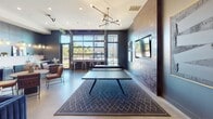 Located at 143 E White St in Rock Hill, SC, this single-floor amenity lounge is designed with a modern industrial aesthetic that blends bold style with functional recreation. The space is anchored by deep navy-blue walls, polished concrete floors, and a dramatic oversized photographic mural depicting industrial machinery, lending the interior a distinctive, urban character.

The main area features a regulation ping pong table set atop a geometric patterned area rug, positioned beneath a sculptural multi-arm pendant light fixture. Large floor-to-ceiling windows and an oversized garage-style door flood the space with natural light and provide views of an outdoor area, creating a seamless indoor-outdoor connection.

A dedicated lounge zone offers channeled navy velvet seating, cognac leather accent chairs, and a shuffleboard table, all set against the striking industrial mural wall. A wall-mounted television is positioned nearby, alongside a small beverage and snack bar area with open shelving. An additional seating cluster with a round table provides a more intimate setting. Curated artwork and layered lighting complete the sophisticated, community-focused ambiance of this well-appointed gathering space.
