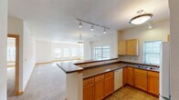 This well-designed single-floor residence offers a thoughtfully laid-out open-concept floor plan that seamlessly connects the kitchen and living areas. The kitchen is appointed with warm honey-toned maple cabinetry, dark laminate countertops, a double stainless steel sink, a built-in dishwasher, and white appliances. A wrap-around peninsula with a dark countertop serves as both a functional workspace and a natural divider between the kitchen and the expansive living area. Track lighting and a decorative pendant fixture illuminate the kitchen, while a large pendant light anchors the transition into the living space. The living area is generously proportioned, featuring plush neutral carpeting, warm wood baseboards, and an abundance of natural light streaming through multiple windows. A dedicated sun-filled nook with a bank of windows along two walls creates an especially bright and inviting corner of the space. The bedrooms are quiet and comfortable, each featuring carpeted floors, recessed window sills, and ample natural light. The bathroom includes a wide vanity with extended countertop storage, tile flooring, and direct access to a walk-in closet with a built-in rod and shelving. Throughout the space, neutral tones and clean lines create a calm, livable atmosphere.