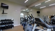 This fitness room is designed to support a well-rounded cardio workout in a compact, functional setting. The space is outfitted with multiple pieces of cardio equipment, including elliptical trainers and at least one treadmill, arranged to make efficient use of the available floor space. Large floor-to-ceiling mirrors line one wall, visually expanding the room and allowing users to monitor their form during exercise. The mirrored wall also reflects an arched window, suggesting natural light enters the space, contributing to a brighter, more open atmosphere. Overhead fluorescent strip lighting ensures the room remains well-lit throughout the day. A wall-mounted flat-screen television is positioned for easy viewing from the cardio machines, adding an entertainment option during workouts. The flooring appears to be carpeted, providing some cushioning underfoot. The neutral, beige-toned walls and clean layout give the space a straightforward, no-frills aesthetic focused on functionality. A dumbbell rack is also visible along one wall, indicating that light free weight training is also accommodated within the room.