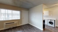 This single-floor space offers a practical and efficiently designed layout that maximizes its available footprint. The main living area features durable dark laminate wood flooring that runs throughout, providing a clean and modern aesthetic. Neutral beige and white walls keep the space feeling bright and versatile, complemented by a textured popcorn ceiling overhead. A window fitted with horizontal blinds allows natural light to filter into the living area, creating a welcoming atmosphere. A wall-mounted HVAC unit is positioned beneath the window, ensuring climate control throughout the seasons. The front entry door is centrally located and framed cleanly against the wall, making for a straightforward and accessible entryway. A half-wall partition separates the living area from the adjacent kitchen space, which is visible to the right and features white cabinetry and standard appliances, including a dark dishwasher or refrigerator unit. The kitchen benefits from its own natural light source, adding to the overall brightness of the space. The layout is open and functional, making it well-suited for straightforward day-to-day living. Clean lines, simple finishes, and a cohesive color palette throughout give this space a tidy and move-in-ready presentation.