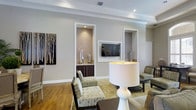 This single-floor space presents a polished and well-appointed lounge environment designed with both function and aesthetics in mind. The interior is defined by a warm neutral color palette — soft greige walls paired with crisp white crown molding and trim — creating a refined, welcoming atmosphere throughout.

A standout architectural feature is the pair of recessed wall niches framed in white, each serving a distinct purpose. One niche houses a wall-mounted flat-screen television, while the adjacent niche functions as a wet bar or display alcove, complete with dark cabinetry and elegant glass lantern accents. These built-in elements add structural character and visual depth to the main wall.

Hardwood flooring in a warm honey tone runs throughout the space, grounding the room with a sense of warmth and continuity. A patterned area rug defines the central seating zone, where upholstered accent chairs in subtle printed fabric are arranged around low dark-wood tables. A prominent white ceramic table lamp with a drum shade anchors the foreground, adding a decorative focal point.

Recessed ceiling lighting provides even, ambient illumination, complemented by the clean lines of the tray-detailed ceiling. The overall design is cohesive and purposeful, reflecting a community or hospitality-oriented space designed to accommodate gathering and relaxation in a tasteful, contemporary setting.