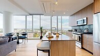 This contemporary 2-bedroom, 1-bathroom residence at 50 Clinton Pl in New Rochelle, NY offers a thoughtfully designed single-floor layout with an emphasis on natural light and panoramic views. Floor-to-ceiling windows span the living and kitchen areas, framing breathtaking vistas of the New Rochelle skyline and Long Island Sound waterfront beyond.

The open-concept kitchen is a standout, featuring a large wood-toned island with seating, stainless steel appliances including a gas range and French door refrigerator, sleek flat-panel cabinetry, and a tile backsplash — all bathed in abundant natural light from the expansive glazing.

The living room is bright and airy, with light hardwood flooring that runs throughout the unit, offering corner window views of the surrounding cityscape. Both bedrooms are well-proportioned; the primary bedroom features large windows with waterfront views, while the second bedroom looks out onto the urban skyline. All rooms benefit from the clean, minimalist aesthetic and warm wood tones consistent throughout the home.

The bathroom is finished with wood-grain vanity cabinetry, stone-tiled walls, a deep soaking tub, and a large backlit mirror, conveying a calm, spa-like atmosphere. The overall design is modern and sophisticated, with a cohesive palette of warm neutrals and natural materials.