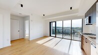 This single-floor residence showcases a clean, contemporary design throughout, with light-toned hardwood flooring and crisp white walls creating a bright, cohesive aesthetic. The open living area is a standout feature, anchored by expansive floor-to-ceiling sliding glass doors that lead to a private balcony, flooding the space with natural light and offering elevated views of the surrounding urban and tree-lined landscape.

The kitchen is thoughtfully designed with a two-tone cabinetry scheme, pairing warm wood lower cabinets with matte dark upper cabinets. Stainless steel appliances, including a French door refrigerator and dishwasher, provide modern functionality alongside a clean subway tile backsplash and undermount sink.

The bedrooms are well-proportioned, each benefiting from black-framed windows that frame treetop and neighborhood views while continuing the unit's modern design language. One bedroom features a dedicated walk-in closet with wrap-around rod storage and matching hardwood flooring.

The bathroom is finished with large-format marble-look floor tiles and a fully glass-enclosed walk-in shower with mosaic tile flooring, a ceiling-mounted rain showerhead, and matte black fixtures — delivering a refined, spa-like atmosphere. Track spotlights and recessed lighting complete the modern, polished interior.