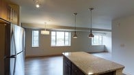 This single-floor space showcases a thoughtfully designed open-concept layout that seamlessly connects the kitchen and living areas. The kitchen is anchored by a substantial island topped with speckled granite countertops and fitted with dark cabinetry, offering both prep space and visual contrast. Three stylish pendant lights hang above the island — two sleek metallic drop pendants and one softer drum shade — providing focused task lighting while adding a contemporary design touch. Additional track lighting is mounted along the ceiling near the kitchen, ensuring excellent illumination throughout. The open living area beyond is generously proportioned, featuring warm hardwood flooring that runs throughout the space. Multiple large windows line the walls, inviting an abundance of natural light and offering pleasant views of the surrounding greenery. The neutral gray wall tones complement the clean, modern aesthetic and provide a versatile backdrop. The overall ambiance is airy and welcoming, with a cohesive design language that balances functionality with modern style — making this an ideal space for both everyday living and entertaining.