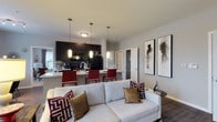 Located at 7600 Huntington Park Dr in Columbus, Ohio, this single-floor residence offers a thoughtfully designed open-concept layout that seamlessly connects the living and kitchen areas. The living space is anchored by a plush white sofa accented with bold red and gold decorative pillows, creating a warm yet contemporary atmosphere. Curated wall art adds a sophisticated touch to the neutral gray walls, contributing to a polished, modern aesthetic throughout. The kitchen is a standout feature, showcasing dark espresso cabinetry, a classic tile backsplash, and sleek black appliances. A breakfast bar with light stone-toned countertops and red bar seating serves as both a functional dining spot and a visual centerpiece. Pendant lighting with warm-toned shades illuminates the kitchen and bar area, enhancing the inviting ambiance. The open design allows natural light to filter through a window near the kitchen, brightening the entire space. Dark hardwood-style flooring runs throughout, providing a rich contrast to the lighter walls and furnishings. A hallway visible from the main living area suggests additional rooms and closet space, completing this well-appointed single-level home in Columbus.