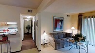 This single-floor residence offers a welcoming and well-organized layout designed for comfortable everyday living. The living area serves as the heart of the space, featuring plush, patterned upholstered seating arranged around a sleek side table and a glass-top accent table. Warm ambient lighting is provided by a classic arc floor lamp and an elegant table lamp, creating a cozy and inviting atmosphere. A large framed vintage-style art print adds a decorative focal point to the wall, lending the room a touch of personality and character. The flooring transitions from soft carpet in the living area to hardwood-style flooring along the hallway, adding visual contrast and warmth. The extended hallway connects the various rooms of the residence, lined with dark wood door frames and accented by additional wall art. At the far end of the corridor, a decorative potted plant introduces a natural element, enhancing the sense of life and warmth throughout the space. The ceilings feature subtle geometric beam detailing and integrated ventilation, adding architectural interest overhead. Recessed and flush-mount lighting fixtures illuminate the hallway evenly. The overall ambiance is calm, neutral, and functional, with a classic residential aesthetic that feels both lived-in and well-maintained.