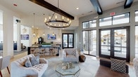 This single-story space is designed with a sophisticated blend of modern and transitional aesthetics, offering a welcoming and well-appointed environment throughout. The main lounge area immediately draws the eye upward to soaring ceilings accented by bold, dark wood beam detailing. A large wrought-iron wagon wheel chandelier with Edison-style bulbs serves as a dramatic focal point, casting warm, ambient light across the space. Plush tufted sofas and curved accent chairs in neutral tones are arranged around a geometric glass coffee table, creating an inviting seating area anchored by a soft area rug. A floor-to-ceiling tiled fireplace surround with a sleek linear gas insert and wall-mounted television adds both warmth and visual interest to one wall. Expansive black-framed glass doors and transom windows flood the interior with natural light while providing direct views to an outdoor area. An adjacent bar-style counter with ornate stools and pendant lighting serves as a casual refreshment station. A separate dining nook features a round table with side chairs alongside original artwork, while the entry area features vaulted ceilings, navy accent walls, and another striking chandelier above light hardwood flooring, making for a grand first impression.