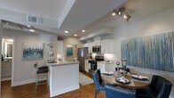 This single-story space showcases an open-concept layout that seamlessly connects the kitchen and dining areas, creating a cohesive and functional living environment. The kitchen is equipped with stainless steel appliances, including a French door refrigerator, gas range, and built-in microwave, all complemented by crisp white cabinetry and light granite countertops. A peninsula island with bar seating provides additional prep space and a casual dining option, while recessed lighting and a tray ceiling detail above add architectural interest.

The warm hardwood flooring flows throughout, grounding the space and providing visual continuity between the kitchen and dining areas. The dining area accommodates a round wooden table paired with upholstered navy blue accent chairs featuring nailhead trim, adding a refined, contemporary touch. Track lighting overhead highlights the dining space effectively.

Neutral gray walls serve as a versatile backdrop, accented by colorful artwork that introduces pops of blue and green throughout. The overall ambiance is modern and polished, balancing practical functionality with thoughtful design details that make the space both livable and visually appealing.
