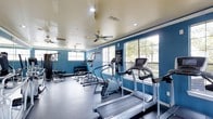 This fitness center offers a comprehensive workout environment designed to support a wide range of exercise routines. The space is immediately recognizable by its bold blue walls, which create an energizing atmosphere conducive to physical activity. Large multi-pane windows line the walls, flooding the interior with generous natural light while providing views of the surrounding greenery outdoors.

The cardio section is well-appointed, featuring multiple treadmills, an elliptical trainer, and a stationary exercise bike, all arranged to take advantage of the natural light and outdoor views. Along the far wall, a dedicated strength training area includes a dumbbell rack with an organized selection of free weights, as well as cable and resistance machines suitable for a full-body workout.

Overhead, ceiling fans are strategically positioned throughout the space to promote air circulation, complementing the HVAC system for year-round comfort. Recessed lighting supplements the natural light during evening hours. The flooring is finished in a durable, dark rubberized material designed to withstand heavy equipment and high foot traffic. An informational board is mounted on the wall, providing guidelines and instructions for facility use. Overall, the space is functional, well-maintained, and thoughtfully laid out.