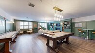 Located at 28 Everett Rd Ext in Albany, NY, this thoughtfully designed single-floor community lounge offers a generous, open layout ideal for both leisure and socializing. The space is defined by rich hardwood flooring and striking sage-green wall paneling that lends a cohesive, upscale aesthetic throughout.

At the heart of the room sits a substantial wooden pool table, illuminated by a brass-finished pendant chandelier with geometric glass globes that serve as a focal point from above. Adjacent to it, a full-length shuffleboard table adds to the recreational appeal of the space.

The living area is anchored by an impressive built-in wall unit finished in teal-green, incorporating open shelving, beadboard cabinetry, a wall-mounted television, and a fireplace flanked by stacked firewood displays — creating a warm, lodge-inspired atmosphere. Comfortable seating, including plush sofas and swivel lounge chairs, is arranged to encourage conversation and relaxation.

Large windows allow abundant natural light to fill the room, with linen drapes softening the look. A bistro-style high-top table with bar stools provides a more casual seating option near the windows. A wet bar area is also visible toward the rear of the space. The overall ambiance is inviting, polished, and well-appointed — a refined gathering space designed for community living.