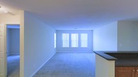 This single-story space presents a straightforward and functional layout with a clean, neutral aesthetic throughout. The interior is characterized by light-toned walls, wall-to-wall carpeting in a soft neutral shade, and white baseboards that provide a cohesive finish across the rooms.

The main living area is an open, rectangular room featuring a set of three large windows dressed with vertical blinds, allowing an abundance of natural light to fill the space. A partial countertop visible at the edge of the frame suggests an adjacent kitchen or bar area, indicating a practical, open-concept flow between living and dining or kitchen spaces.

A second room, likely a bedroom or additional living space, mirrors the minimal design with multiple windows fitted with vertical blinds spanning two walls, creating a bright and airy atmosphere. The textured ceiling with a subtle popcorn finish adds a modest architectural detail to this room.

Both spaces are unfurnished, allowing the full scale of each room to be appreciated. The overall ambiance is calm and neutral, providing a blank canvas well-suited for customization.