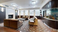 This elegantly designed multi-use space offers a seamlessly connected layout across a generously sized single floor. The main lounge area is anchored by a built-in fireplace with a wall-mounted flat-screen television, complemented by plush seating arrangements set atop a distinctive geometric-patterned area rug over rich hardwood flooring. Large windows along the perimeter flood the space with abundant natural light while offering views of the surrounding urban skyline.

A striking centerpiece aquarium, framed in dark cabinetry, serves as a dramatic divider between the lounge and a dedicated reading nook. The library area features glass shelving stocked with books, positioned alongside the windows to take advantage of natural light.

The kitchen and bar area is outfitted with a white quartz countertop island, mosaic tile detailing, bar seating, stainless steel appliances, a wine cooler, and stylish cylindrical pendant lighting. Adjacent to the kitchen is a formal conference or dining room with a large dark wood table, upholstered seating, recessed lighting, and built-in AV cabinetry, framed by frosted glass double doors. The space combines contemporary design with warm tones throughout for a polished, professional ambiance.