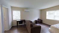 This single-floor space offers a clean, functional layout designed for comfortable everyday living. The main living area is finished with rich, dark hardwood flooring that adds warmth and character throughout. Neutral, light-toned walls create a bright and open atmosphere, complemented by natural light streaming in through a window that frames views of surrounding greenery.

Climate comfort is handled by a wall-mounted mini-split air conditioning and heating unit, providing efficient year-round temperature control. The room accommodates a plush, L-shaped sofa that serves as the centerpiece of the living space, alongside a wooden desk and chair positioned near the window — ideal for work or study. A dark wood media console with glass-panel doors provides additional storage and display space, completing the functional furnishings of the room.

The ceiling follows a subtle angled pitch toward one end of the room, adding a modest architectural detail to the otherwise straightforward interior. Baseboards and trim are finished in white, offering a clean contrast against the darker flooring. Overall, the space presents a modest yet practical environment, well-suited for individuals seeking a simple, self-contained living arrangement with essential amenities and a quiet, nature-adjacent feel.