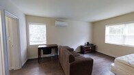 This single-floor space offers a clean, functional layout designed for comfortable everyday living. The main living area is finished with rich, dark hardwood flooring that adds warmth and character throughout. Neutral, light-toned walls create a bright and open atmosphere, complemented by natural light streaming in through a window that frames views of surrounding greenery.

Climate comfort is handled by a wall-mounted mini-split air conditioning and heating unit, providing efficient year-round temperature control. The room accommodates a plush, L-shaped sofa that serves as the centerpiece of the living space, alongside a wooden desk and chair positioned near the window — ideal for work or study. A dark wood media console with glass-panel doors provides additional storage and display space, completing the functional furnishings of the room.

The ceiling follows a subtle angled pitch toward one end of the room, adding a modest architectural detail to the otherwise straightforward interior. Baseboards and trim are finished in white, offering a clean contrast against the darker flooring. Overall, the space presents a modest yet practical environment, well-suited for individuals seeking a simple, self-contained living arrangement with essential amenities and a quiet, nature-adjacent feel.
