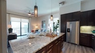 This single-floor residence showcases a thoughtfully designed open-concept layout that seamlessly connects the kitchen and living areas. The kitchen serves as the centerpiece of the home, anchored by a generous island topped with striking granite countertops featuring intricate natural veining. Rich, dark-toned cabinetry with sleek hardware provides ample storage, while stainless steel appliances, including a full-size French door refrigerator, add a polished, modern touch. Pendant lighting with decorative crystal accents hangs above the island, casting warm, ambient light throughout the space. The living area flows naturally from the kitchen, bathed in natural light streaming through large windows dressed with layered roman shades and curtains. A ceiling fan with integrated lighting adds both comfort and style to the living space, which is tastefully furnished with a neutral-toned sofa and colorful accent pieces. Warm wood-look flooring runs throughout, unifying the open floor plan. A private balcony accessible from the living area offers expansive views, bringing the outdoors in and enhancing the airy feel of this well-appointed residence. The overall ambiance is modern, warm, and inviting.