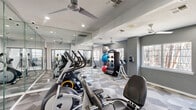 Located at 2700 Central Drive in Bedford, TX, this fitness center is organized across two dedicated exercise rooms on a single level, offering a comprehensive workout environment. The space features a distinctive geometric-patterned carpet flooring in shades of gray and white throughout, lending a modern, energetic aesthetic to the interior.

The main exercise room is outfitted with a variety of cardio equipment, including elliptical trainers, treadmills, and stationary bikes. Large floor-to-ceiling mirrors line one wall, visually expanding the space and supporting proper form during workouts. A rack of medicine balls and exercise balls adds functional variety for strength and flexibility training, while a dumbbell rack provides additional free-weight options. Ceiling fans and recessed lighting ensure comfortable, well-lit conditions throughout.

The second exercise room is dedicated to cycling and additional cardio, housing multiple indoor cycling bikes and treadmills. Natural light filters in through several windows fitted with blinds, creating a bright and motivating atmosphere. A ceiling fan maintains airflow, complementing the room's clean, light gray walls and textured ceiling.

The facility's layout and equipment selection cater to a broad range of fitness needs, from cardio and cycling to strength and functional training, making it a versatile and practical workout destination.