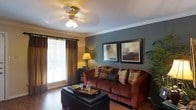 This single-floor space offers a warm and inviting atmosphere, highlighted by rich hardwood flooring and elegant crown molding that frame each room with classic architectural detail. The living area serves as the focal point of the home, featuring a deep, plush sofa in warm burgundy tones accented by an array of decorative throw pillows in earthy hues. A leather ottoman coffee table anchors the seating arrangement, providing both style and functionality. Two framed art pieces adorn a slate-blue accent wall, adding depth and visual interest to the room. Natural light filters in through a large window dressed with layered curtains — sheer blinds paired with rich gold and brown draping panels — creating a cozy yet bright ambiance. A classic ceiling fan with a light fixture provides both airflow and overhead illumination. A warm-toned floor lamp adds a soft glow to complement the natural light. Touches of greenery, including a lush indoor palm, bring life and freshness to the space. The overall aesthetic balances comfort and style, making this a welcoming and well-designed living environment.