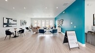 The Resident Lounge at 2115 Park Springs Circle in Arlington, Texas, is a thoughtfully designed community gathering space that combines contemporary style with welcoming functionality. The single-floor layout flows openly, making excellent use of its generous square footage to create distinct yet connected zones for socializing, lounging, and working.

A striking bold teal accent wall anchors the space visually, contrasting beautifully against the crisp white ceilings and light gray walls throughout. Light wood-look flooring runs continuously across the entire area, lending warmth and cohesion to the open layout. Recessed lighting and a ceiling fan ensure the space is well-lit and comfortable throughout the day.

The lounge area is furnished with stylish navy blue accent chairs featuring abstract-print throw pillows, arranged around a central coffee table to encourage conversation. A plush sectional sofa anchors a secondary seating zone, while decorative metallic wall art adds an artistic focal point against the teal wall. Tall decorative vases and candle accents contribute to the curated, upscale ambiance.

A dedicated dining area features multiple round white tables paired with modern black chairs, ideal for casual meals or small gatherings. Abstract artwork adorns the walls in this section, adding a gallery-like quality. Large windows throughout the lounge flood the space with natural light and offer views of the outdoor pool area, seamlessly connecting the interior to the surrounding community.