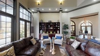 Located at 5555 Long Prairie Trace in Richmond, TX, this single-story community space offers an impressive and inviting atmosphere defined by upscale architectural details and thoughtful design. The grand entrance foyer greets visitors with slate tile flooring, soaring ceilings adorned with dark crown molding, and arched doorways that lend a refined, classic character throughout.

The main gathering area is a showpiece, anchored by a dramatic floor-to-ceiling stacked stone fireplace and flanked by expansive, dark-framed grid windows that flood the space with abundant natural light and offer views of the surrounding outdoor areas. Rich leather sofas, patterned accent chairs, and layered area rugs create a warm, inviting seating arrangement ideal for relaxed gatherings.

A prominent built-in bookcase with rich mahogany tones provides both display and storage, adding depth and warmth to the living area. Coffered ceiling details with dark wood trim accents further elevate the space's sophisticated aesthetic. Structural columns define zones within the open layout while maintaining a seamless visual flow between areas.

Adjacent rooms, accessible through arched and glass-paned doorways, offer additional functional spaces suited for meetings or quiet activities. Throughout, a cohesive palette of warm neutrals, natural stone, and deep wood tones creates a welcoming, polished ambiance well-suited for community gathering and daily interaction.