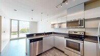Located in Seattle's Capitol Hill neighborhood, this modern one-bedroom, one-bathroom residence offers a well-designed single-floor layout with clean contemporary finishes throughout. The open-concept kitchen and living area serves as the heart of the home, featuring sleek white cabinetry extending to the ceiling, dark countertops, and a full suite of stainless steel appliances including an electric range, built-in microwave, dishwasher, and refrigerator. Stylish pendant lighting accents the kitchen, which flows into the living area and is anchored by warm wood-look flooring. Large floor-to-ceiling windows flood the space with natural light and frame views of the surrounding Seattle streetscape and greenery. The bedroom offers a comfortable footprint with a built-in closet featuring natural wood-finish sliding doors, wood-look flooring, and access to ample natural light through generous windows with neighborhood views. A baseboard heater ensures year-round comfort. The bathroom is finished with speckled tile flooring, a full bathtub, and in-unit washer/dryer hookups, providing practical functionality. Throughout the residence, natural wood-tone doors add warmth against the neutral palette, creating a cohesive and modern urban living environment.