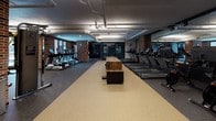 Located at 601 Meeting Street in Charleston, South Carolina, this well-appointed fitness center offers a comprehensive, single-floor facility designed to support a full range of workout routines. The space is characterized by a striking industrial aesthetic, with exposed brick columns, overhead ductwork, and a combination of rubber and hard-surface flooring that defines the different functional zones.

The main cardio area is expansive, featuring a long row of TRUE treadmills flanked by elliptical trainers and stationary cycling bikes. Large floor-to-ceiling windows along one wall flood the space with natural light and provide views of the surrounding Charleston streetscape. Full-length mirrors along the opposite wall visually expand the room and aid in form monitoring. A rustic wood-topped bench anchors the center of the cardio floor.

Adjacent to the cardio zone is a dedicated strength training area equipped with a cable machine, adjustable bench, medicine balls, and a tiered kettlebell rack, all set on durable dark rubber flooring. Exposed brick columns frame the space and contribute to the industrial-chic character of the facility.

A separate studio room offers a bright, open environment with light wood flooring, a ceiling fan, recessed and track lighting, yoga mats, stability balls, resistance tools, and a wall-mounted mirror with a barre — ideal for yoga, stretching, or group fitness. Access to the facility is provided through sleek glass-panel doors with black metal framing.