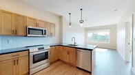 This thoughtfully designed single-floor unit showcases a contemporary aesthetic throughout. The open kitchen is a standout feature, equipped with stainless steel appliances including a refrigerator, range, microwave, and dishwasher. Warm natural wood cabinetry pairs elegantly with dark countertops, while a light blue subway tile backsplash adds a subtle pop of color. Pendant lighting with an industrial-modern style hangs above the kitchen peninsula, which also features an integrated sink — an ideal setup for both cooking and casual dining. The kitchen flows naturally toward a bright living area framed by large windows that invite abundant natural light and offer views of a colorful urban mural, providing a vibrant and artistic backdrop. Hardwood-style flooring runs throughout the living and bedroom areas, contributing to a cohesive, clean look. The bedroom is generously sized, bathed in natural light from a large picture window, and includes a built-in closet with sliding doors for ample storage. The bathroom features slate-toned tile flooring, a rich wood vanity with a crisp white countertop, brushed nickel fixtures, and a frameless mirror with medicine cabinet. Overall, the space offers a clean, modern ambiance well-suited for everyday living.