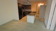 This single-floor residence is designed with an open-concept layout that seamlessly connects the kitchen and living areas, creating a spacious and practical environment. The kitchen is a standout feature, showcasing rich dark wood cabinetry that extends to the upper walls, providing generous storage. Complementing the cabinetry are sleek dark granite-style countertops, a black refrigerator, black range, and a built-in microwave, giving the kitchen a cohesive, contemporary aesthetic. A central kitchen island with a light base and matching dark countertop serves as both a prep space and a casual gathering point, anchoring the room. Track lighting overhead illuminates the kitchen and flows into the adjacent hallway, ensuring the space is well-lit throughout. The transition from tile flooring in the kitchen to soft carpeting in the living area creates a natural delineation between the two spaces while maintaining an open feel. A hallway leading further into the residence hints at additional rooms beyond, with a paneled door visible in the background. The overall ambiance is warm and inviting, with a neutral color palette that offers a versatile backdrop for a variety of interior styles.