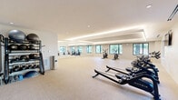 Located at 57 Main St in Marlborough, MA, this well-appointed fitness center offers a thoughtfully designed single-floor exercise space suited for a comprehensive workout experience. The layout is divided into distinct functional zones, including a dedicated cardio area, a free weights and accessories section, and an open floor space ideal for stretching or functional training.

The cardio area is equipped with a row of Matrix rowing machines and stationary cycling bikes, all positioned to face large, black-framed windows that frame views of the surrounding greenery and outdoor environment. Natural light flows generously throughout, creating an energizing atmosphere. A wall-mounted flat-screen TV provides added convenience for users during workouts.

A well-organized free weights station features a multi-tier rack stocked with kettlebells, medicine balls, stability balls, and resistance accessories. Sleek glass-panel sliding doors separate additional sections of the facility, lending a modern, open feel to the overall design.

The interior is finished with neutral-toned carpeting, crisp white walls, and a tray ceiling accented by elegant indirect cove lighting and recessed downlights. The clean, contemporary aesthetic is complemented by full-length mirrors that visually expand the space. The fitness center presents a polished, professional environment well-suited for daily exercise routines.