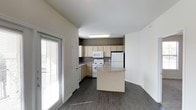 This single-floor space offers a clean, functional layout centered around an open-concept kitchen and living area. The kitchen is thoughtfully designed with light wood cabinetry that provides ample storage, white appliances including a range, over-the-range microwave, and refrigerator, and a freestanding center island with a laminate countertop that serves as both a workspace and casual dining spot. The cabinetry is set against a warm taupe accent wall, adding a subtle contrast to the otherwise neutral white walls and creating a defined kitchen zone within the open floor plan. Dark hardwood-style flooring runs throughout the space, lending a sense of warmth and cohesion. Natural light filters in generously through a sliding glass door flanked by a full-length window with blinds, brightening the interior and providing a connection to the outdoors. The layout flows openly between the kitchen and adjacent living area, making the space feel welcoming and easy to navigate. Flush-mounted ceiling lighting ensures the kitchen remains well-illuminated. The overall ambiance is clean and contemporary, with a neutral palette that offers a versatile backdrop for a variety of furnishing styles.