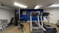 Located at 9978 Guilford Rd in Jessup, Maryland, this single-floor fitness center offers a well-organized and functional workout environment designed to support a range of exercise routines. The space is finished with neutral gray walls, durable carpet tile flooring throughout, and bright overhead fluorescent and recessed lighting that keeps the facility well-illuminated.

The main workout area is divided into distinct zones. A dedicated cardio section houses multiple treadmills and an elliptical trainer, positioned near windows that allow natural light to filter in from outside. A bold navy blue accent wall anchors the strength training area, which is equipped with a multi-station cable machine, a weight bench, and additional strength apparatus. Framed artwork on the accent wall adds a touch of visual interest to the workout environment.

An open multi-purpose area provides additional floor space for stretching or functional exercises, complemented by a freestanding coat rack and a bulletin board mounted on the wall. A wall-mounted television is also present for entertainment during workouts.

Supporting amenities include a water cooler station conveniently located near a dedicated restroom with ADA-compliant signage, ensuring accessibility for all users. A secondary door provides direct access to the exterior, enhancing convenience and ventilation. Overall, this fitness center offers a clean, practical, and comfortable environment for everyday exercise.