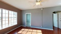 This single-story space presents a clean and functional layout with a cohesive interior design throughout. The main room immediately draws attention with its rich hardwood flooring, which catches natural light streaming in through a multi-pane window, casting a warm grid of sunlight across the floor. The walls are finished in a cool, neutral grey tone, complemented by crisp white baseboards and door trim that provide a sharp contrast and a polished look. A ceiling fan with an integrated light fixture is centrally mounted, offering both air circulation and ambient lighting. Crown molding runs along the upper edges of the walls, adding a subtle architectural detail that elevates the room's character. A doorway leads into an adjoining room, which features lighter wall tones and continues the carpeted or smooth flooring, suggesting a versatile secondary space. The overall ambiance is calm and understated, with the interplay of natural light and neutral finishes creating a welcoming, move-in-ready environment. The layout offers practical connectivity between rooms while maintaining a sense of openness and simplicity throughout the space.