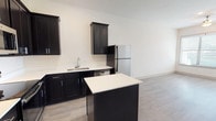 This single-floor, 1-bedroom, 1-bathroom unit offers a thoughtfully designed layout with modern finishes throughout. The kitchen serves as a standout feature, showcasing bold dark shaker-style cabinetry contrasted against crisp white countertops and a full suite of stainless steel appliances, including a refrigerator, range, microwave, and dishwasher. A central kitchen island provides additional prep and counter space, while recessed and track lighting illuminate the area effectively. Light-toned wood-style flooring runs throughout the unit, creating a cohesive and clean aesthetic. The open layout connects the kitchen to the main living area, maximizing the sense of space. The bedroom is bright and airy, benefiting from a large window that invites natural light and features a ceiling fan for added comfort. A generously sized walk-in closet with built-in shelving and dual hanging rods offers excellent storage and organization potential. The bathroom is finished with classic white subway tile surrounding the soaking tub and shower combination, complemented by neutral tile flooring and chrome fixtures. Clean white walls and consistent flooring throughout the unit contribute to a bright, modern, and move-in-ready ambiance.