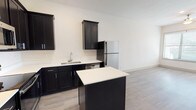 This single-floor, 1-bedroom, 1-bathroom unit offers a thoughtfully designed layout with modern finishes throughout. The kitchen serves as a standout feature, showcasing bold dark shaker-style cabinetry contrasted against crisp white countertops and a full suite of stainless steel appliances, including a refrigerator, range, microwave, and dishwasher. A central kitchen island provides additional prep and counter space, while recessed and track lighting illuminate the area effectively. Light-toned wood-style flooring runs throughout the unit, creating a cohesive and clean aesthetic. The open layout connects the kitchen to the main living area, maximizing the sense of space. The bedroom is bright and airy, benefiting from a large window that invites natural light and features a ceiling fan for added comfort. A generously sized walk-in closet with built-in shelving and dual hanging rods offers excellent storage and organization potential. The bathroom is finished with classic white subway tile surrounding the soaking tub and shower combination, complemented by neutral tile flooring and chrome fixtures. Clean white walls and consistent flooring throughout the unit contribute to a bright, modern, and move-in-ready ambiance.